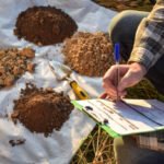 A man writes on a clipboard while examining soil, illustrating a geotechnical soil investigation process.
